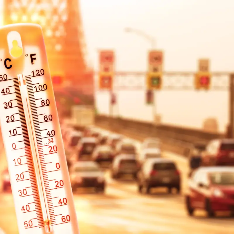 A thermometer showing a high temperature in front of a heat-hazed highway filled with cars.