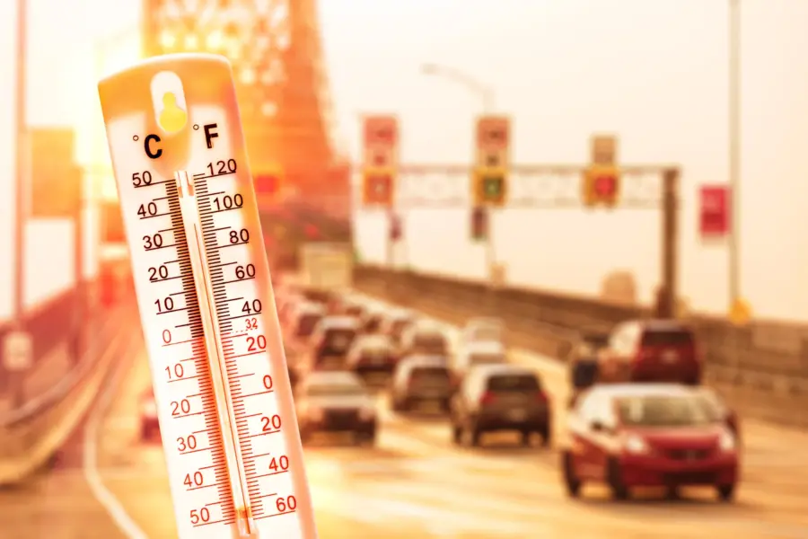 A thermometer showing a high temperature in front of a heat-hazed highway filled with cars.